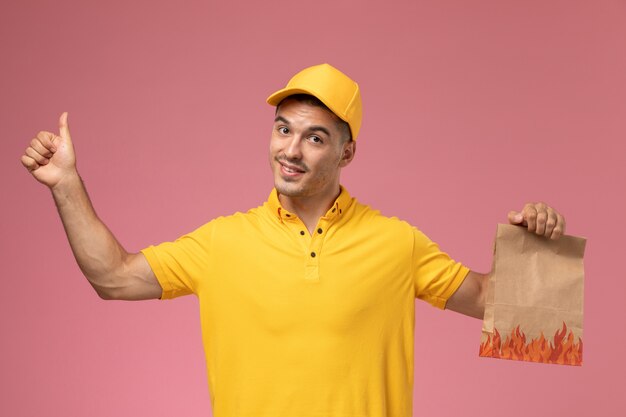 Courier masculino de uniforme amarelo sorrindo e segurando um pacote de comida no fundo rosa