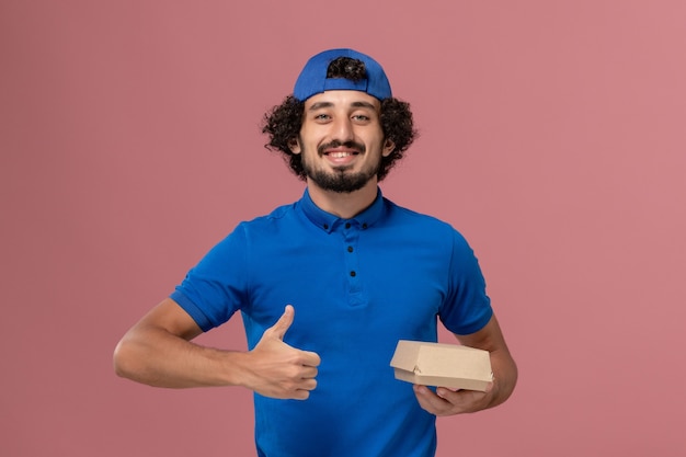 Courier masculino de uniforme azul e capa segurando um pequeno pacote de entrega de comida na parede rosa. empregado de empresa de serviço de entrega uniforme