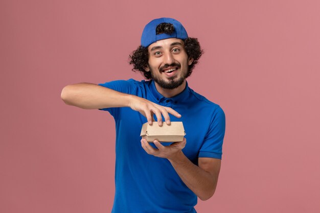 Courier masculino de uniforme azul e capa segurando um pequeno pacote de entrega de comida na parede rosa funcionário da empresa de entrega de uniforme