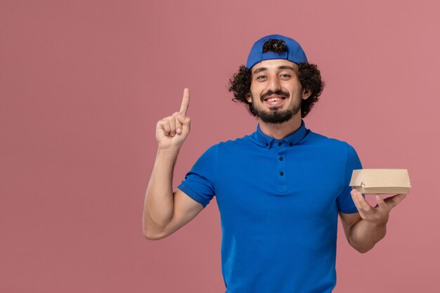 Courier masculino de uniforme azul e capa segurando um pequeno pacote de entrega de comida na parede rosa