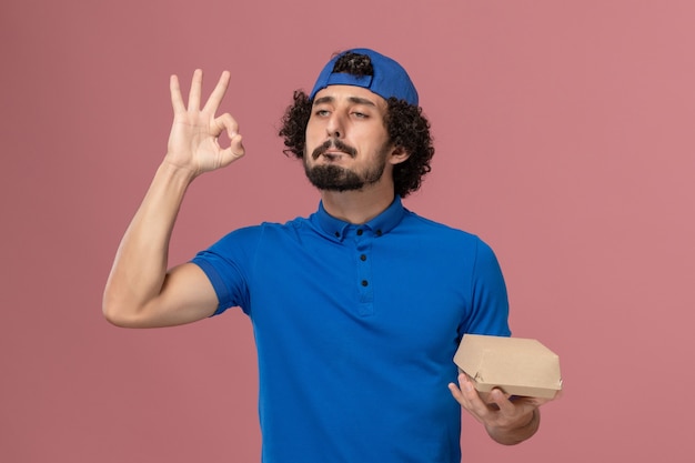 Courier masculino de uniforme azul e capa segurando um pequeno pacote de entrega de comida na parede rosa