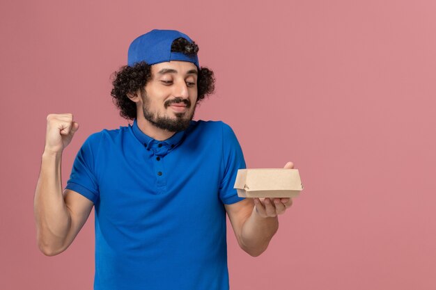 Courier masculino de uniforme azul e capa segurando um pequeno pacote de entrega de comida na parede rosa