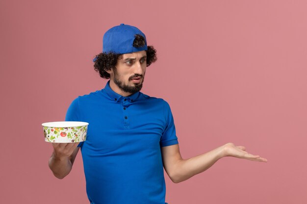 Courier masculino de uniforme azul e capa segurando uma tigela redonda na parede rosa