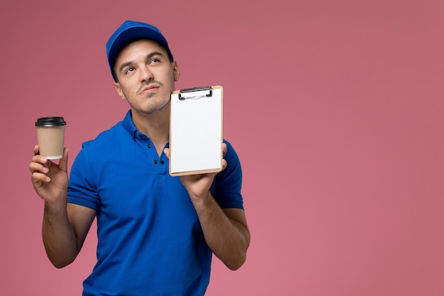 Courier masculino de uniforme azul segurando um bloco de notas e café na parede rosa, entrega de serviço de uniforme de trabalhador