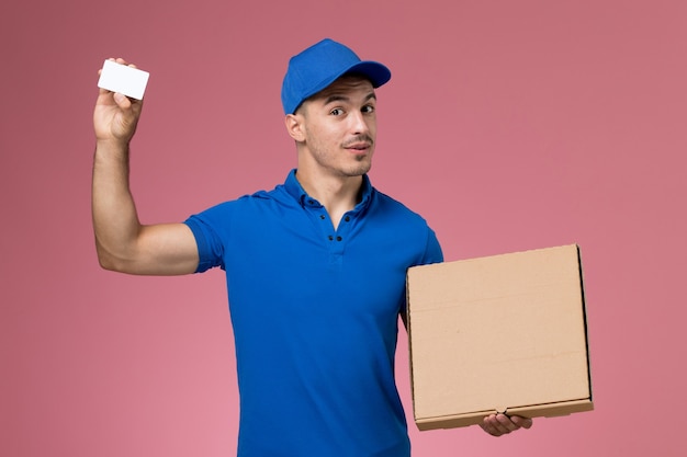 Courier masculino de uniforme azul segurando uma caixa de comida com cartão na parede rosa, entrega de serviço de uniforme de trabalhador