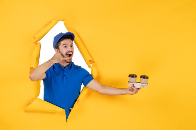 Courier masculino de uniforme azul segurando xícaras de café no chão amarelo.