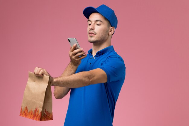 Courier masculino de uniforme azul tirando uma foto de pacote de comida na parede rosa, entrega de serviço de trabalhador uniforme
