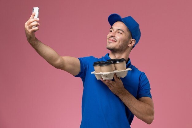 Courier masculino de uniforme azul tirando uma selfie com entrega de xícaras de café na parede rosa, serviço de trabalho uniforme.