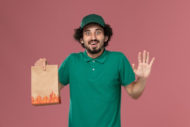 Courier masculino de uniforme verde e capa segurando um pacote de comida de papel na mesa rosa serviço de entrega uniforme.