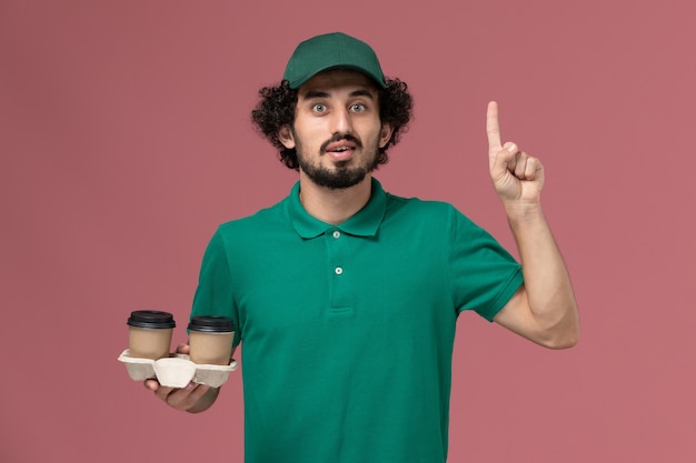 Courier masculino de uniforme verde e capa segurando xícaras de café no fundo rosa. Serviço de entrega uniforme de trabalho