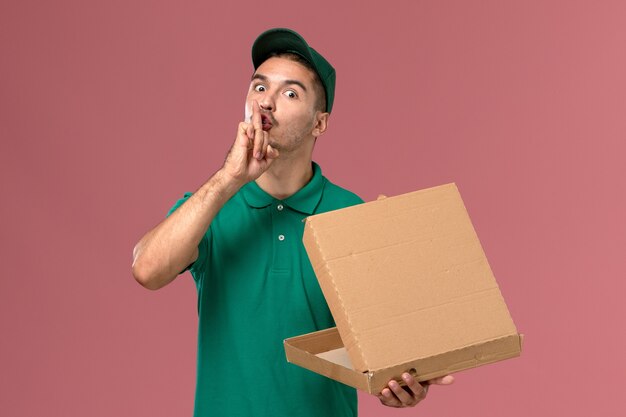 Courier masculino de uniforme verde segurando a caixa de comida e abrindo-a em fundo rosa