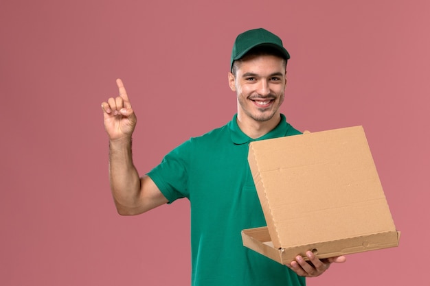 Courier masculino de uniforme verde segurando a caixa de comida e abrindo-a em um fundo rosa claro