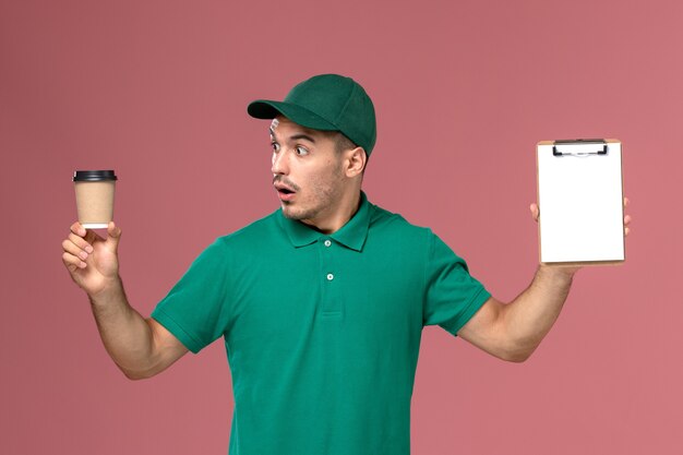 Courier masculino de uniforme verde segurando a xícara de café de entrega e o bloco de notas com expressão de surpresa na mesa rosa