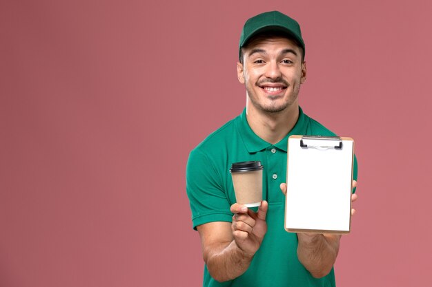 Foto grátis courier masculino de uniforme verde segurando a xícara de café de entrega e o bloco de notas com um sorriso na mesa rosa claro