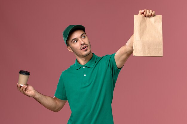 Courier masculino de uniforme verde segurando a xícara de café de entrega e o pacote de comida no fundo rosa claro