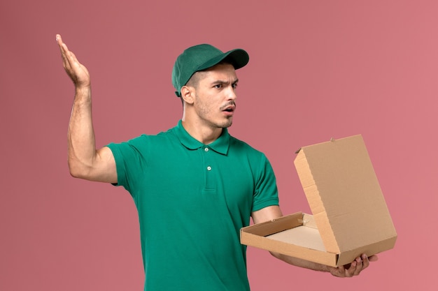 Courier masculino de uniforme verde segurando e abrindo a caixa de comida com expressão de descontentamento no fundo rosa