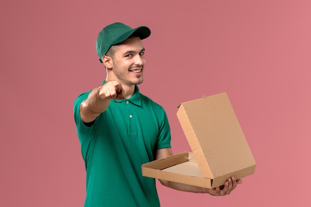 Courier masculino de uniforme verde segurando e abrindo a caixa de comida no fundo rosa claro