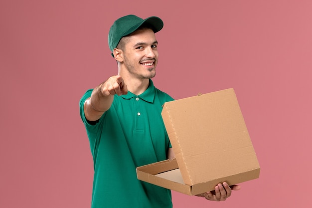 Courier masculino de uniforme verde segurando e abrindo a caixa de comida no fundo rosa claro