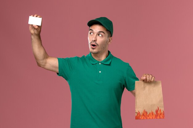 Courier masculino de uniforme verde segurando o pacote de comida e o cartão na mesa rosa