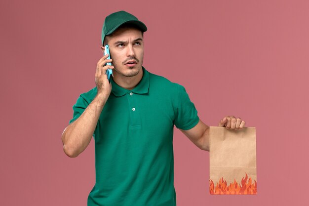 Courier masculino de uniforme verde segurando um pacote de comida enquanto fala ao telefone sobre fundo rosa