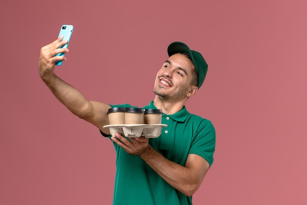 Courier masculino de uniforme verde segurando xícaras de café marrons e tirando uma foto com eles na mesa rosa