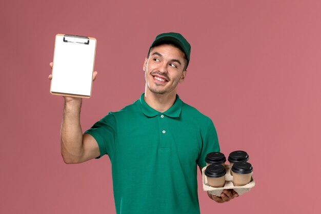Courier masculino de uniforme verde segurando xícaras de café marrons e um bloco de notas com um sorriso no fundo rosa