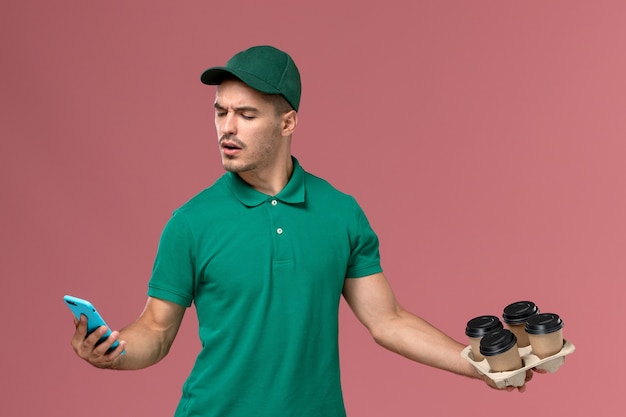 Courier masculino de uniforme verde segurando xícaras de café marrons usando o telefone no fundo rosa.