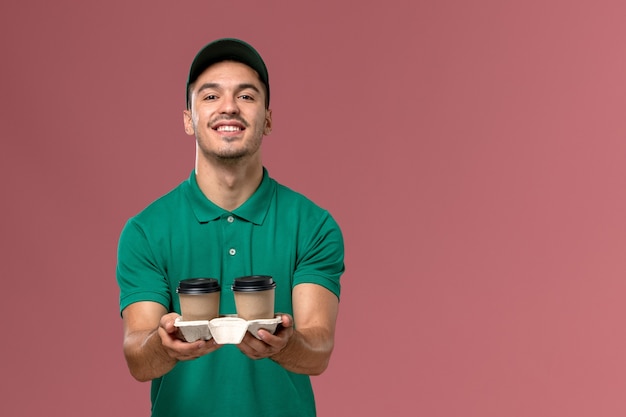 Courier masculino de uniforme verde segurando xícaras de café sorrindo no fundo rosa claro