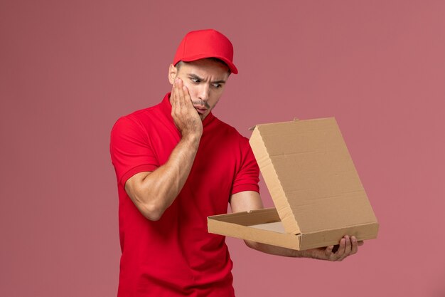 Courier masculino de uniforme vermelho e capa segurando a caixa de comida e abrindo na mesa rosa trabalhador