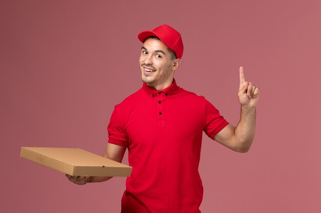 Courier masculino de uniforme vermelho e capa segurando a caixa de entrega de comida na parede rosa