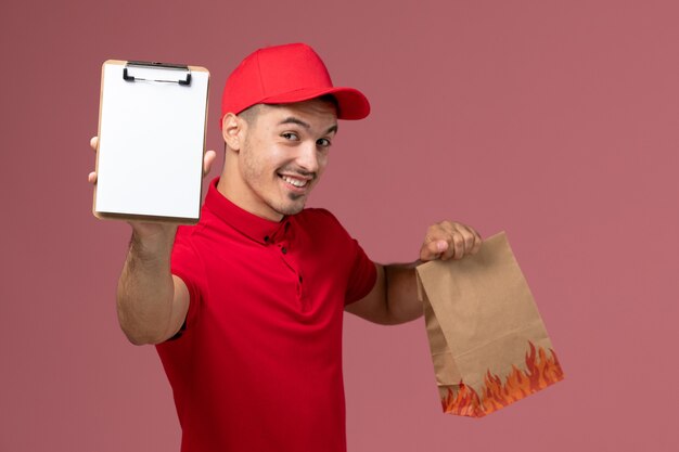 Courier masculino de uniforme vermelho e capa segurando o pacote de comida e o bloco de notas sorrindo na parede rosa