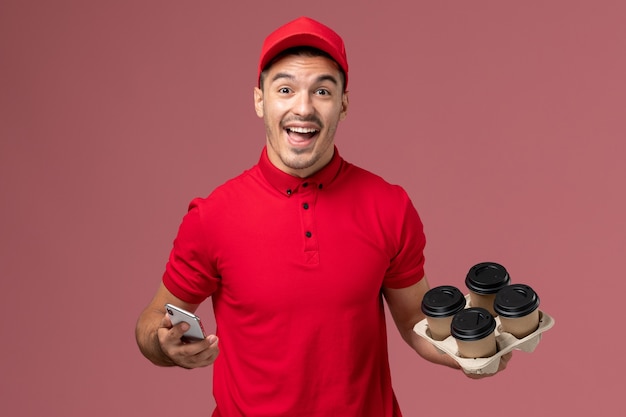 Courier masculino de uniforme vermelho e capa segurando o telefone de entrega de copos de café na parede rosa