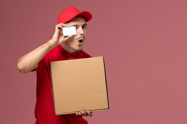 Courier masculino de uniforme vermelho e capa segurando um cartão branco e uma caixa de comida na mesa rosa