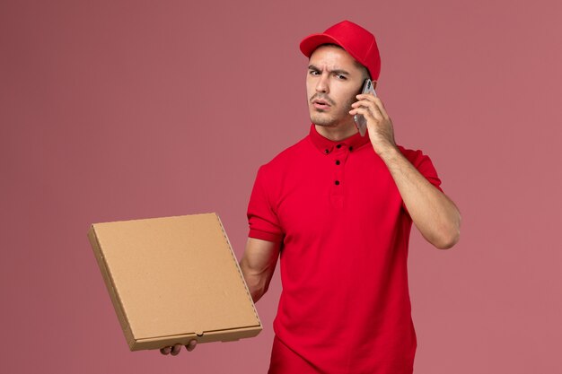 Courier masculino de uniforme vermelho e capa segurando uma caixa de comida falando ao telefone na mesa rosa