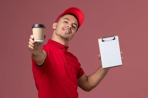 Courier masculino de uniforme vermelho segurando a xícara de café de entrega e o bloco de notas na parede rosa.
