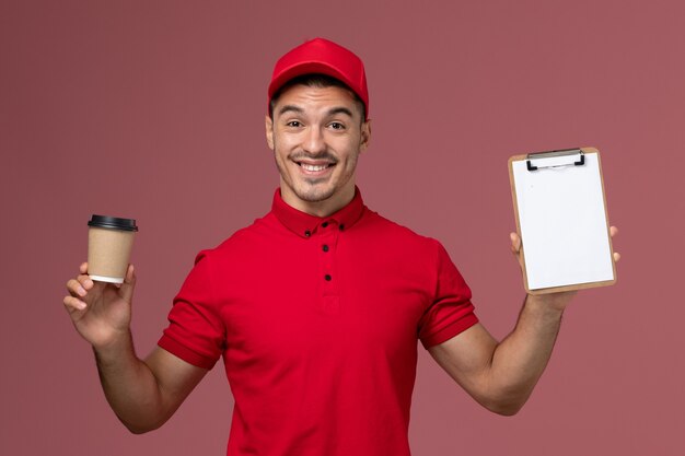 Courier masculino de uniforme vermelho segurando a xícara de café de entrega e o bloco de notas na parede rosa.