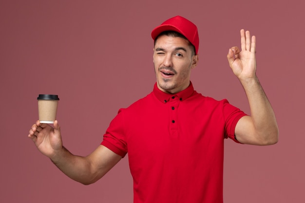 Courier masculino de uniforme vermelho segurando a xícara de café de entrega e piscando na parede rosa trabalhador masculino