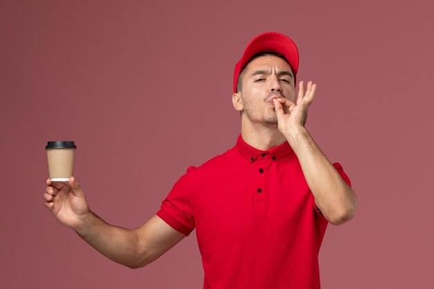 Courier masculino de uniforme vermelho segurando a xícara de café de entrega mostrando sinal saboroso na mesa rosa claro trabalhador masculino