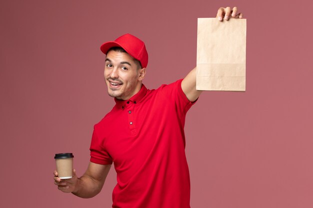 Courier masculino de uniforme vermelho segurando uma xícara de café de entrega e um pacote de comida na parede rosa.