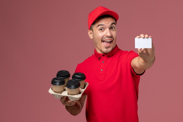 Courier masculino de uniforme vermelho segurando xícaras de café com cartão branco na parede rosa serviço de entrega de trabalho uniforme