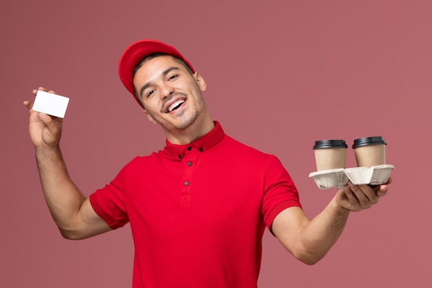 Courier masculino de uniforme vermelho segurando xícaras de café marrom e cartão na parede rosa claro