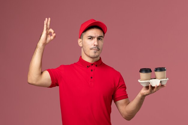 Courier masculino de uniforme vermelho segurando xícaras de café marrom na parede rosa claro trabalhador masculino