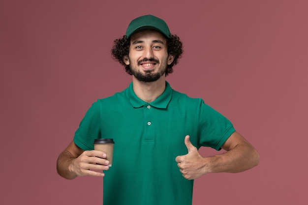 Courier masculino de vista frontal em uniforme verde e capa segurando a xícara de café de entrega sorrindo no trabalho de serviço de entrega uniforme de fundo rosa