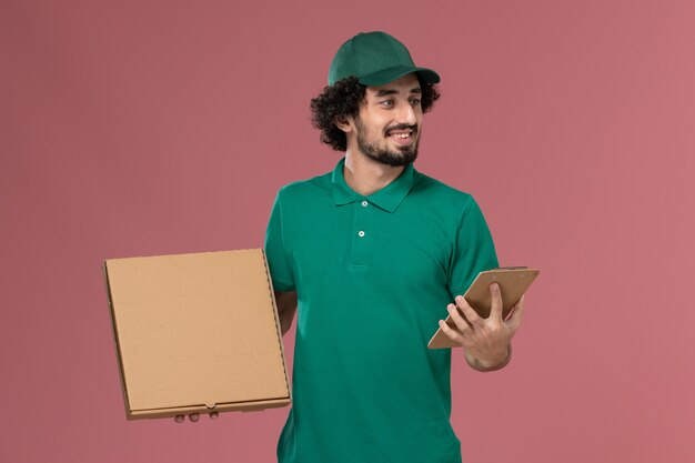 Courier masculino de vista frontal em uniforme verde e capa segurando o bloco de notas de caixa de comida de entrega sorrindo no fundo rosa.