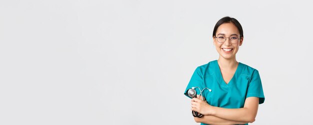 Covid-19, doença de coronavírus, conceito de profissionais de saúde. Médica asiática feliz confiante, médica de óculos, cruzando o peito e sorrindo, segurando o estetoscópio para exame