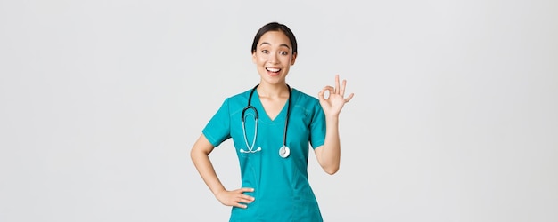 Covid-19, profissionais de saúde, conceito de pandemia. Surpresa e feliz médica asiática, enfermeira de avental mostrando um gesto bem e sorrindo espantada, elogia o bom trabalho, concordo com alguém.