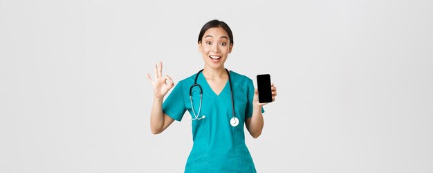 Covid-19, profissionais de saúde e conceito de medicina online. Médica asiática impressionada e satisfeita tentando novo método de consulta on-line durante o coronavírus, mostrando aplicativo de smartphone e gesto bem