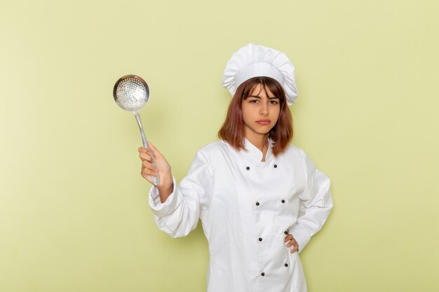 Cozinheira de frente para a cozinheira em um terno branco segurando uma colher de prata grande na superfície verde
