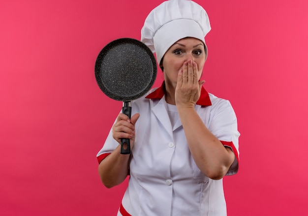 Cozinheira de meia-idade assustada com uniforme de chef segurando uma frigideira tapando a boca com a mão e espaço de cópia