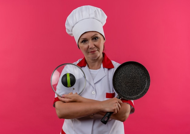 Foto grátis cozinheira de meia-idade com uniforme de chef segurando a frigideira e a tampa na parede rosa isolada com espaço de cópia